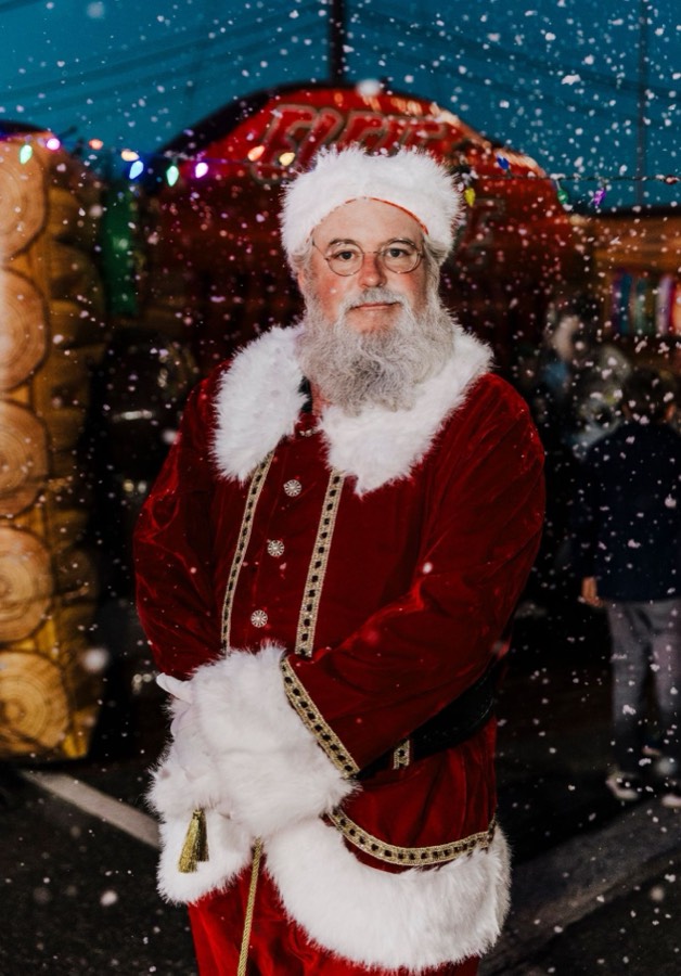 Santa standing in falling snow with holiday lights behind him